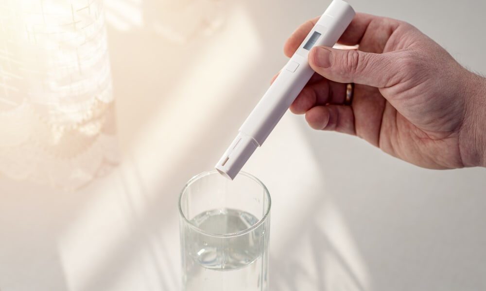 Eine Hand hält ein TDS Messgerät über ein Glas Wasser, um den TDS-Wert (Gesamtgehalt gelöster Feststoffe) zu überprüfen.