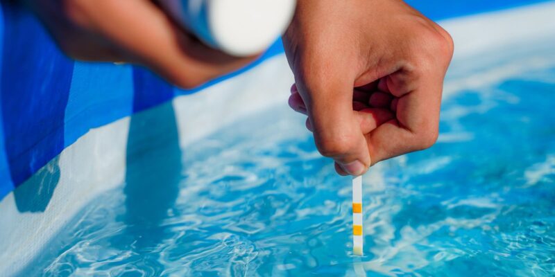 Ein Chlor Teststreifen wird in einen Pool getauscht, die einzelnen Plättchen auf den Teststreifen haben sich gelblich verfärbt, dies gibt eine grobe Einschätzung, wie viel Chlor im Wasser enthalten ist bzw. wie viel Chlor dem Wasser hinzugefügt werden sollte