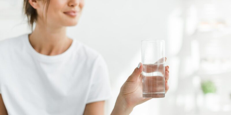 Eine Frau, die ein halbvolles Wasserglas hält, ob in dem Wasser Chlor enthalten ist, ist mit dem bloßen Auge nicht zu erkennen. Wenn man am Wasser riecht und einen Chlorgeruch wahrnimmt, kann dies ein Zeichen für Chlor im Trinkwasser sein