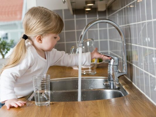 Ein kleines Mädchen mit blonden Haaren und einem Zopf steht an einem Küchenspülbecken und hält eine Hand unter den laufenden Wasserhahn. Neben ihr steht ein Glas mit Wasser. Blei im Trinkwasser schädlich, besonders für Kleinkinder und Säuglinge