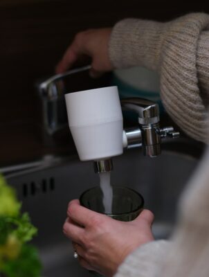Das Bild zeigt eine Person, die einen riva Wasserhahnfilter benutzt, um ein Glas mit Wasser zu füllen. Der Filter ist am Auslauf des Wasserhahns befestigt und sorgt dafür, dass das Wasser gefiltert wird, bevor es ins Glas fließt.