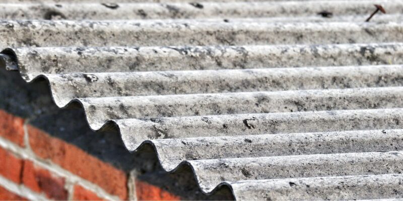 Eine Nahaufnahme von Wellplatten aus Asbestzement, die als Dachabdeckung auf einem Gebäude mit rotem Ziegelstein verwendet werden. Die Oberfläche der Platten wirkt rau und verwittert.